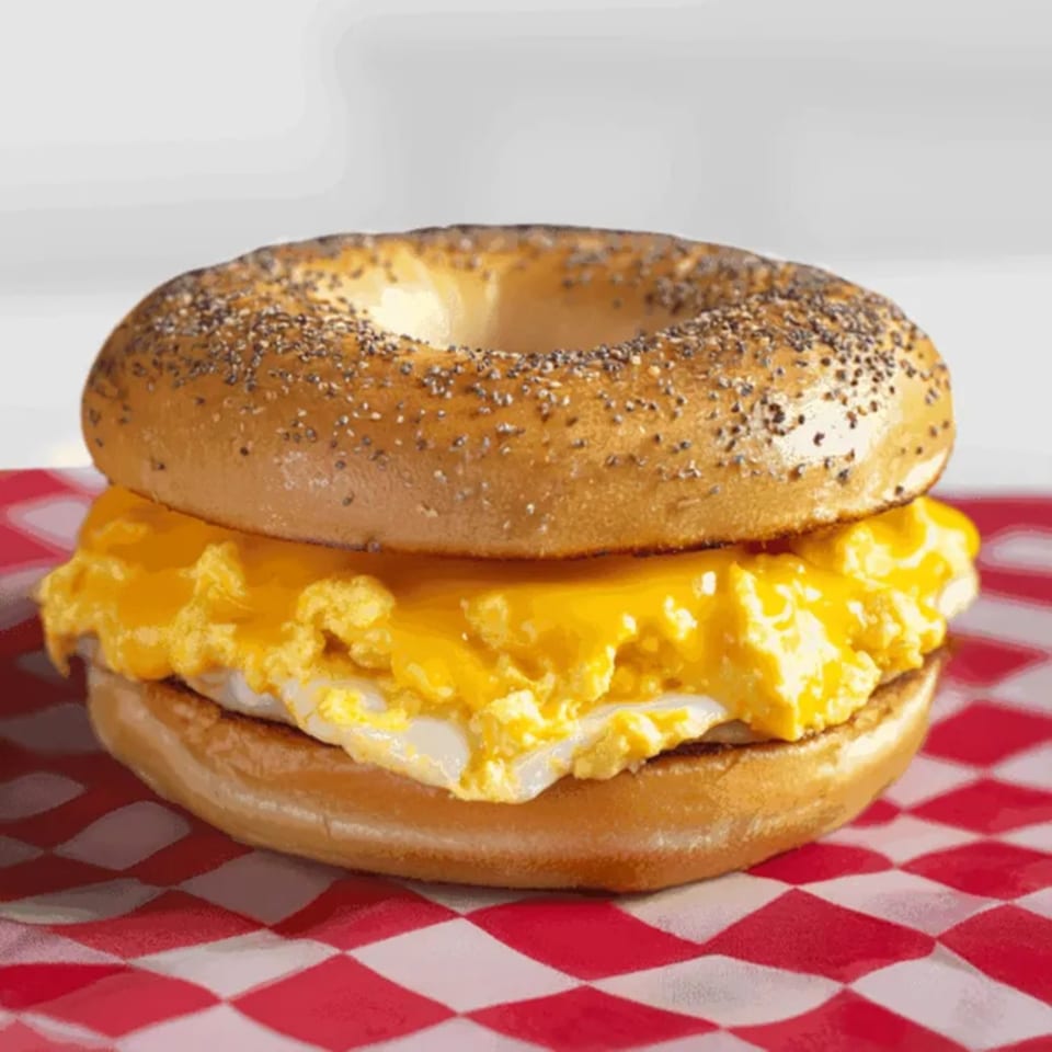 A poppy seed bagel with scrambled eggs and melted cheese on a red and white checkered paper.