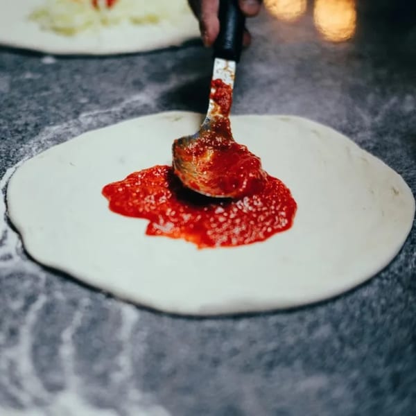 A hand spreads red pizza sauce onto a round, raw Bozzelli's pizza dough.