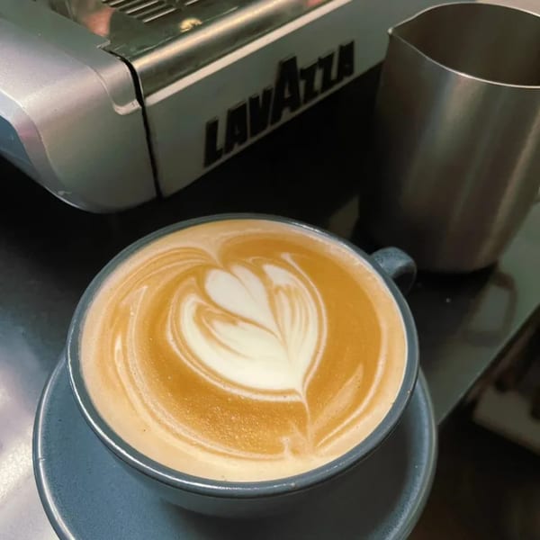 A latte with heart-shaped latte art sits on a saucer next to a Lavazza espresso machine.