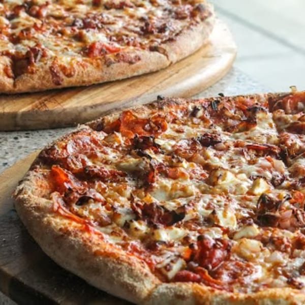 Two Bozzelli's pizzas with melted cheese and various toppings on wooden cutting boards.